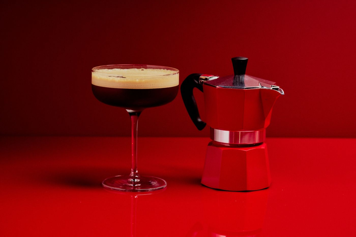 An Espresso Martini next to a coffee pot on a red background  