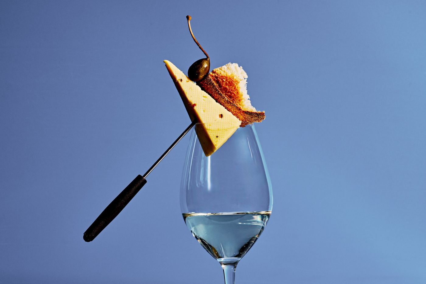 A glass of wine with a skewer holding cheese, bread and a caperberry