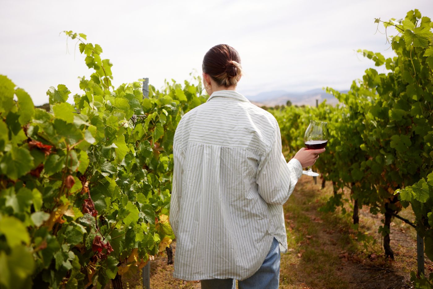 A woman stands between rows of grapevines with a glass of wine