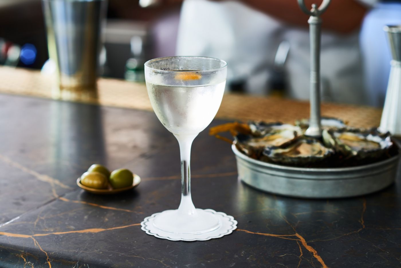 A martini on a bar with olives and oysters in the background 