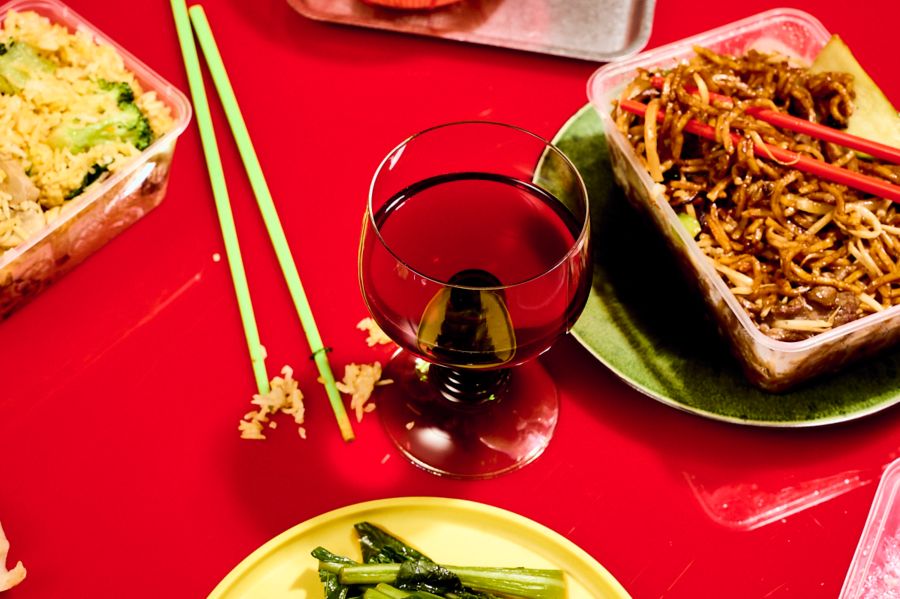 A takeway container of Chinese-style noodles with a glass of red