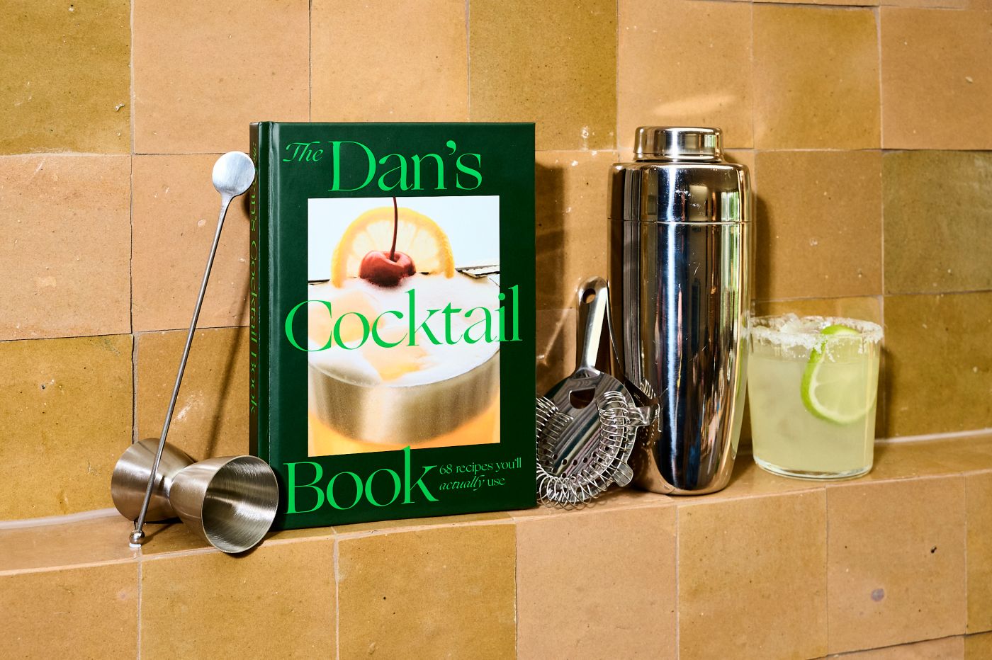 The Dan's Cocktail Book on a shelf with bar tools and a cocktail